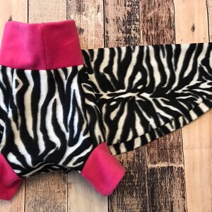 May include: A black and white zebra print dog sweater with pink trim. The sweater has a turtleneck and a long tail.