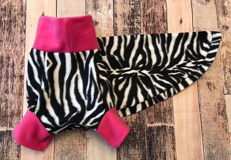 May include: A black and white zebra print dog sweater with pink trim. The sweater has a turtleneck and a long tail.