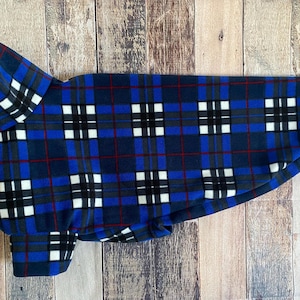 May include: Blue, black, white, and red plaid dog coat. The dog coat has a high collar and a rounded bottom. The plaid pattern is made up of squares and lines. The coat is made of a soft, fleece-like material.