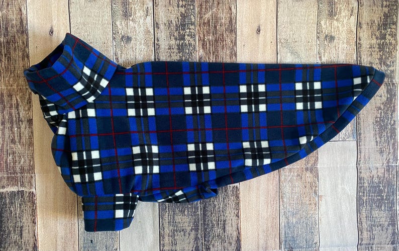 May include: Blue, black, white, and red plaid dog coat. The dog coat has a high collar and a rounded bottom. The plaid pattern is made up of squares and lines. The coat is made of a soft, fleece-like material.