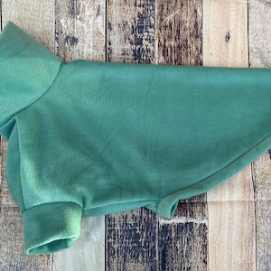 May include: A sage green fleece dog coat with a turtleneck collar and sleeves. The dog apparel is laid flat on a wooden surface. The coat is designed to keep dogs warm and comfortable.