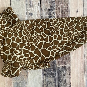 Op de afbeelding: Een bruin en crèmekleurig dierenjasje met giraffenprint. De jas is gemaakt van zacht fleecemateriaal en heeft een hoge kraag en een afgeronde vorm. Het giraffenpatroon bedekt de hele jas, waardoor een unieke en stijlvolle look voor huisdieren ontstaat.