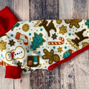 Op de afbeelding: Een witte hondentrui met een rode rand en een kerstthema print met gingerbread men, snoepstokken, rendieren en kerstboom.