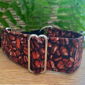 May include: A dog collar featuring a coffee bean design in brown and black hues. It has a silver-coloured metal buckle and fittings. Green foliage and a wooden surface form the background.