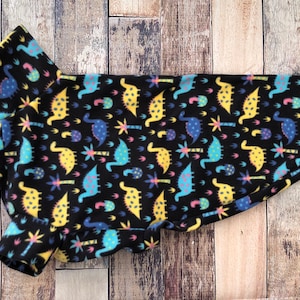 Op de afbeelding: Een zwarte fleece hondentrui met een kleurrijke dinosaurusprint. De dinosaurussen zijn geel, blauw en roze.