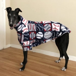 May include: A greyhound dog wearing a baseball-themed fleece jacket. The jacket features a pattern of baseballs in red, white, and grey squares on a navy blue background. The dog is standing on a wooden floor.
