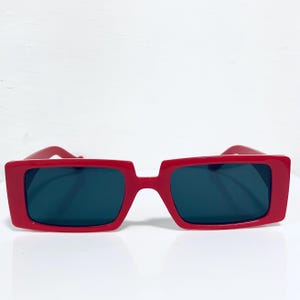 May include: Red rectangular sunglasses with dark lenses. The sunglasses have a bold, modern design with a thick frame. The frames are a vibrant red color, and the lenses are a dark, solid color.
