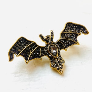 Op de afbeelding: Een goudkleurige vleermuisbroche met donkere, gefacetteerde kristallen op de vleugels en het lichaam. Een enkele, heldere, druppelvormige kristal is in het midden van de borst geplaatst en vormt een blikvanger op de donkere stenen inleg.