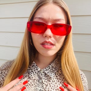 May include: Bright red rectangular sunglasses with a thick frame. The person is wearing a leopard print shirt. The person has long blonde hair and red nail polish. The sunglasses are a bold fashion statement.