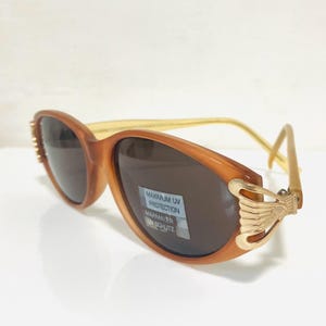 Puede incluir: Gafas de sol ovaladas marrones con lentes oscuras. Las monturas son de color marrón claro con detalles dorados en los laterales. Las lentes tienen una etiqueta "MAXIMUM UV PROTECTION".
