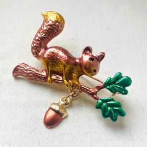 May include: A detailed squirrel brooch in shades of copper, gold, and green. The squirrel is perched on a branch with green leaves and an acorn charm. The brooch is a whimsical accessory.