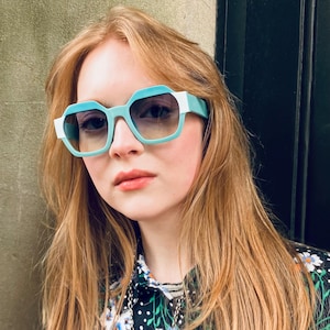 May include: Geometric sunglasses with a light blue and white frame. The lenses are gradient, transitioning from a darker shade at the top to a lighter shade at the bottom. The sunglasses are worn by a person with long, light red hair.