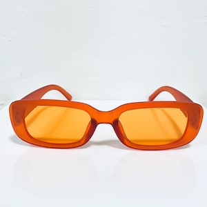 May include: Orange-tinted sunglasses with a rectangular frame. The sunglasses have a solid orange color throughout the frame and lenses. The arms curve slightly at the temples. The sunglasses are set against a white background.