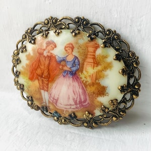 May include: Oval-shaped vintage brooch with a painted scene of a couple in period clothing. The image is set within a filigree gold-tone metal frame. The couple is in a garden setting, with the woman in a pink dress and the man in a brown coat.