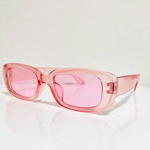 May include: Pink rectangular sunglasses with a translucent frame and lenses. The sunglasses have a retro, oversized design, offering a bold and stylish look. The lenses are a solid pink color.