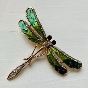 Puede incluir: Broche de libélula dorada con alas de esmalte verde y detalles de cristal transparente. El cuerpo de la libélula presenta una fila de cristales, y las alas están detalladas con diferentes tonos de verde. Accesorio decorativo.