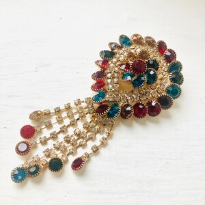 Peut inclure: Une broche en métal doré présentant une monture en forme de cachemire ornée de strass transparents et de bijoux ronds multicolores dans des tons rouge, bleu sarcelle et ambre. La pièce comprend des chaînes pendantes terminées par des pierres.