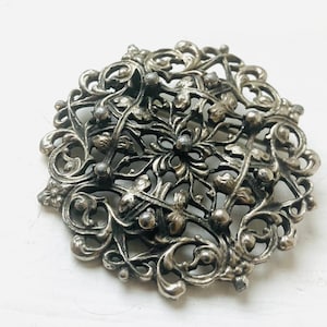Op de afbeelding: Oornamentale zilverkleurige broche met een gedetailleerd bloemen- en krulwerkontwerp. De cirkelvormige broche heeft een centraal bloemmotief omgeven door delicate bladeren en kralenaccenten. Een accessoire in vintage-stijl.