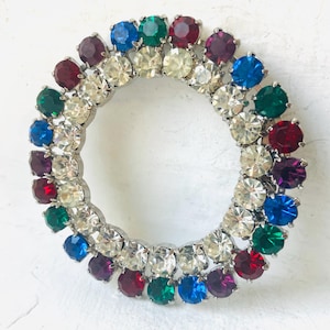 Peut inclure: Une broche circulaire avec un cadre en métal argenté, ornée d'un anneau de pierres précieuses colorées et facettées. Les pierres précieuses sont dans les tons de rouge, vert, bleu, violet et transparent, créant un design vibrant. Ce bijou de style vintage est adapté à diverses occasions.