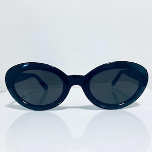 Può includere: Occhiali da sole ovali neri con lenti scure. Gli occhiali hanno una montatura spessa e arrotondata e un design elegante. Le lenti scure offrono ombra e protezione dal sole. Su sfondo bianco.