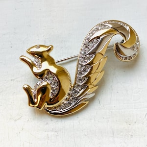May include: A gold-toned squirrel brooch with a silver and crystal-studded tail. The squirrel is posed with its front paws raised, and the brooch has a pin on the back for attachment. The brooch is a vintage piece.