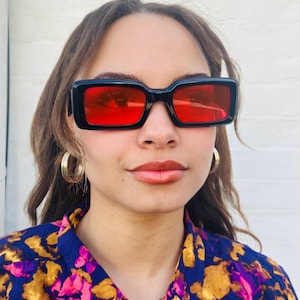 May include: Black rectangular sunglasses with bright red lenses. The sunglasses have a thick frame and are worn by a person. The person is wearing a blue shirt with a floral pattern and gold hoop earrings.