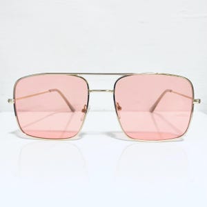 May include: Gold-framed sunglasses with square pink lenses. The sunglasses have a double bridge and a classic aviator style. The lenses are a soft, pastel pink colour, and the frames are a shiny gold. The sunglasses are on a white surface.