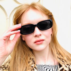 Peut inclure: Lunettes de soleil rectangulaires noires avec une monture épaisse. Les lunettes de soleil sont tenues par une main. La personne porte une veste à imprimé léopard et un haut rayé noir et blanc.