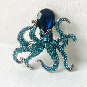 Op de afbeelding: Een broche in de vorm van een octopus met een grote, gefacetteerde donkerblauwe kristallen kop en acht zilverkleurige metalen tentakels ingelegd met talloze kleine, heldere turkooiskleurige strass-steentjes. Inclusief twee kleine zwarte kristallen ogen.