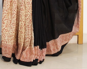 May include: A black and cream cotton saree with a red floral print and a blue and red border. The saree is draped over a wooden chair.