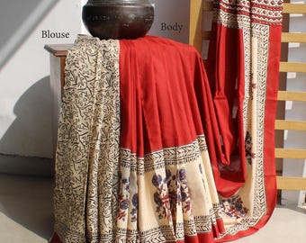 May include: A red and cream-colored saree with floral and geometric patterns. The saree is draped over a wooden ladder and a small table. A dark brown vase sits on the table, and the saree's border features a detailed design.