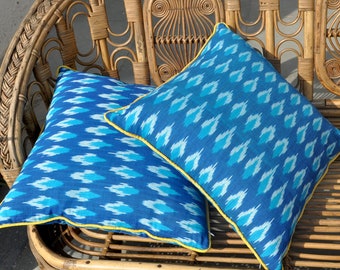 May include: Two blue and white patterned throw pillows with yellow trim are placed on a wicker sofa. The sofa has a woven design and is made of natural materials.