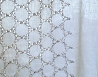 Pode incluir: Uma cortina branca transparente com um delicado padrão bordado de círculos e flores. A cortina está pendurada na frente de uma janela com vista para um prédio ao fundo. O texto "Vedah Designs" é visível no canto inferior direito da imagem.