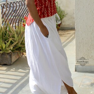 May include: White loose-fitting trousers featuring an elastic waistband with a drawstring, side pockets, and a split hem design. The trousers are paired with a red top featuring a white pattern. The text VedahDesigns is visible.