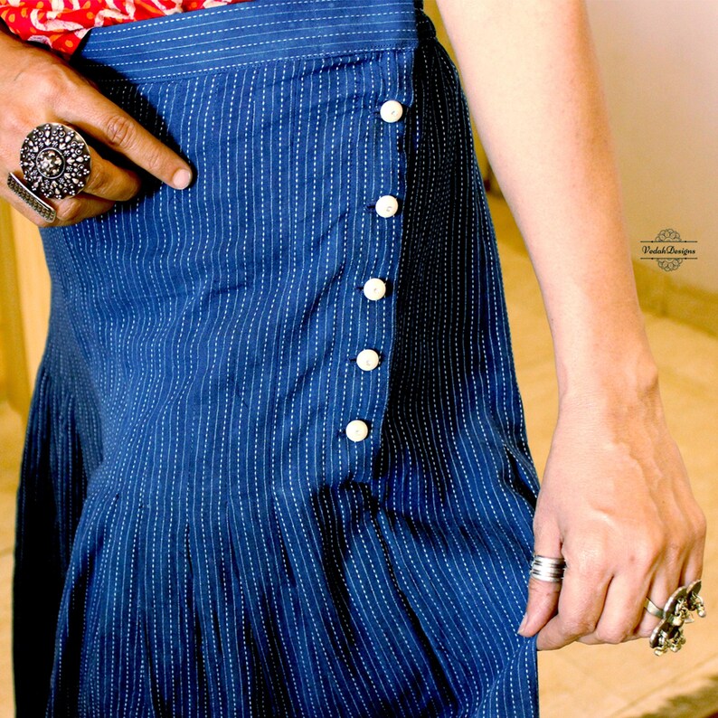 May include: A dark blue skirt featuring a vertical white pinstripe pattern and a side closure with five round white buttons. The garment has a structured waistband and pleated detailing along the lower section, with the text Vedah Designs visible.