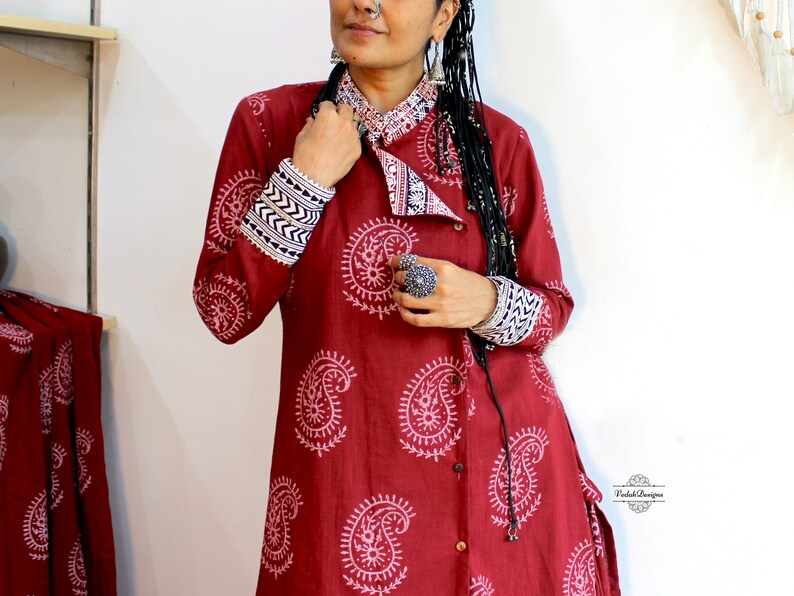 May include: A red long-sleeved tunic with a white paisley print and a contrasting white and blue trim on the cuffs. The tunic has a button-up front and a collar.