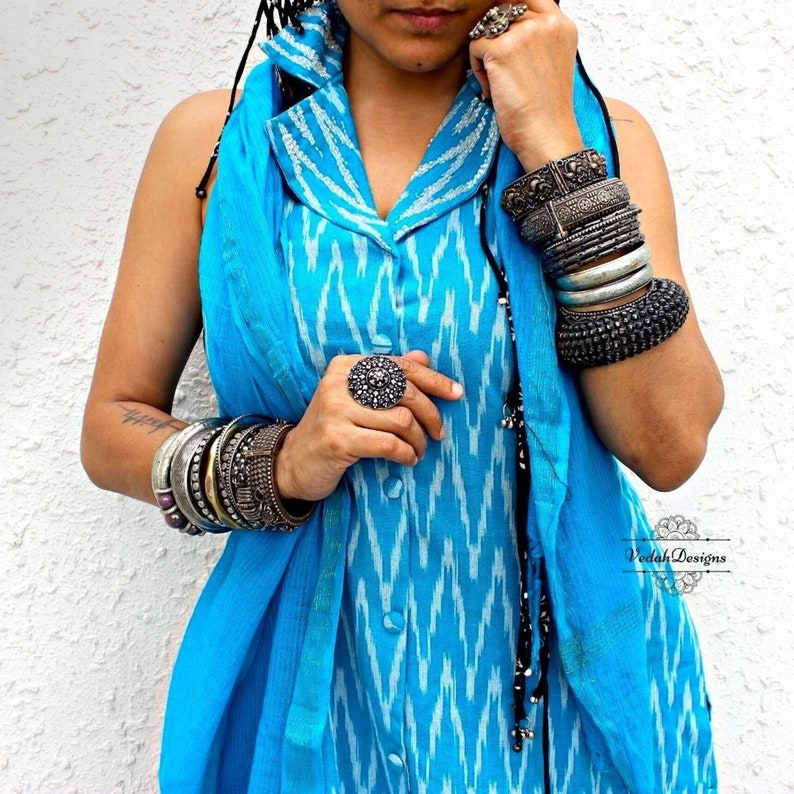 K&ouml;nnte beinhalten: Eine Frau tr&auml;gt ein blaues und wei&szlig;es Ikat-Kleid mit einer schwarzen Perlenkette und silbernen Armreifen. Sie tr&auml;gt einen silbernen Ring mit einem gro&szlig;en Stein an ihrer rechten Hand. Der Text "Vedah Designs" ist in der rechten unteren Ecke des Bildes sichtbar.