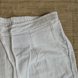 May include: White pants featuring an elasticized waistband with a matching drawstring tie. The garment has a textured fabric surface and includes side pockets. The design includes vertical seam detailing along the front hip area.