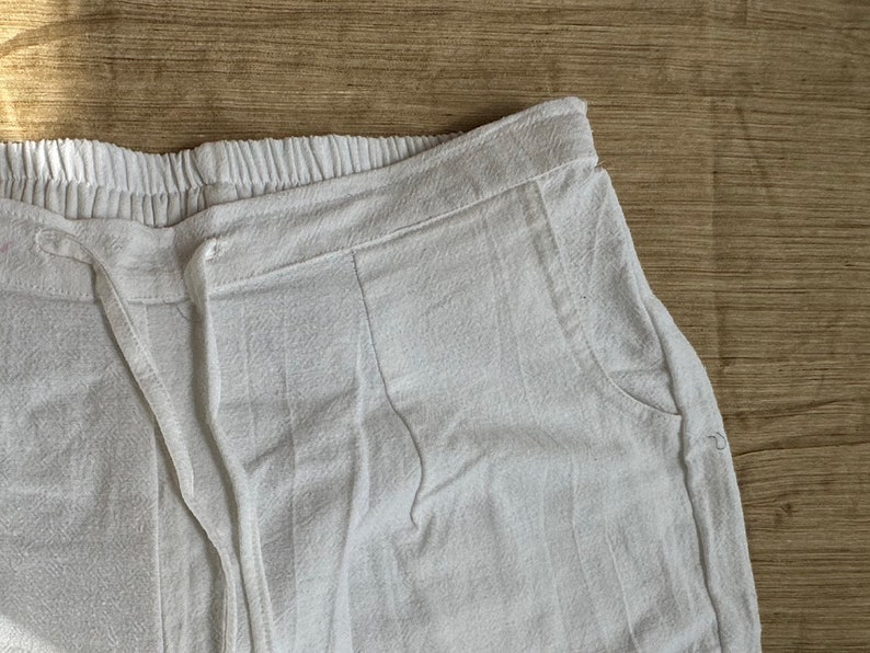 May include: White pants featuring an elasticized waistband with a matching drawstring tie. The garment has a textured fabric surface and includes side pockets. The design includes vertical seam detailing along the front hip area.