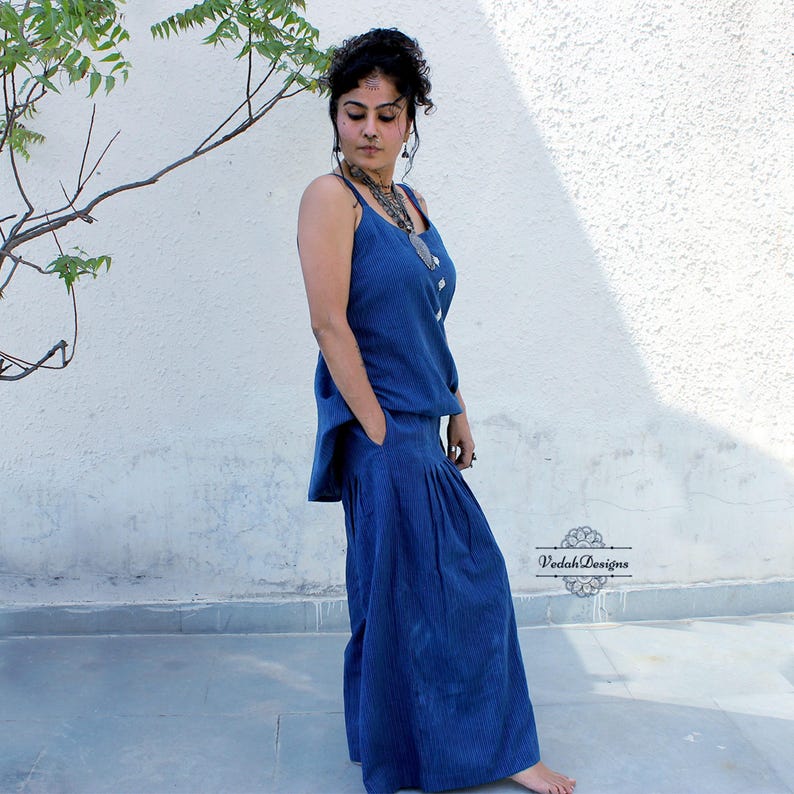 May include: A dark blue sleeveless top and matching wide-leg trousers set featuring a subtle vertical pinstripe pattern. The top has thin shoulder straps and the trousers include side pockets and a pleated waistline. The text Vedah Designs is visible.