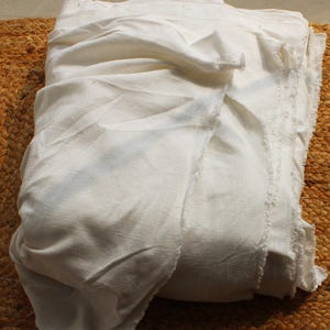 May include: A large piece of off-white, unbleached fabric with a visible weave and raw, frayed edges. The material is folded into multiple layers, creating soft, natural drapes and shadows across the surface of the textile.