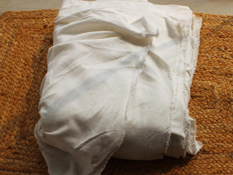 May include: A large piece of off-white, unbleached fabric with a visible weave and raw, frayed edges. The material is folded into multiple layers, creating soft, natural drapes and shadows across the surface of the textile.