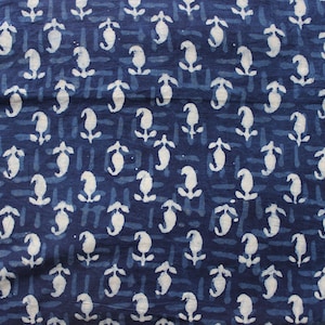 May include: A dark blue fabric featuring a repeating white block-printed pattern of small, stylized floral motifs. The background consists of horizontal rows of short, dashed lines in a slightly lighter shade of blue, creating a textured appearance.