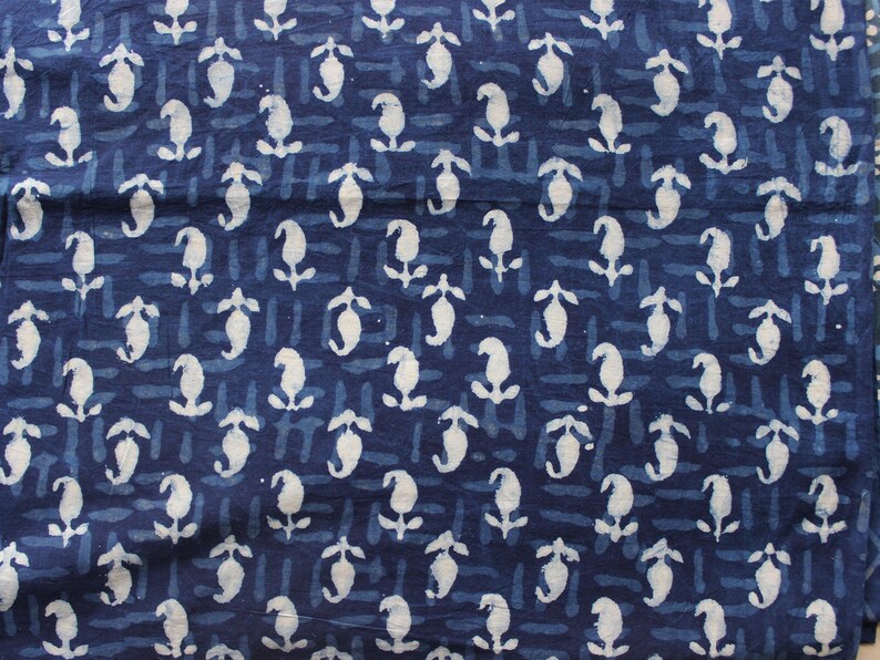 May include: A dark blue fabric featuring a repeating white block-printed pattern of small, stylized floral motifs. The background consists of horizontal rows of short, dashed lines in a slightly lighter shade of blue, creating a textured appearance.