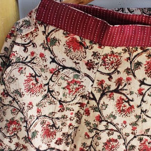 Op de afbeelding: Een rok met een cr&egrave;mekleurige basis en een herhalend bloemenpatroon in tinten rood, zwart en groen. Het kledingstuk heeft een brede, donkerrode tailleband met dunne, verticale witte strepen en zichtbare stiksels over de gehele stof.