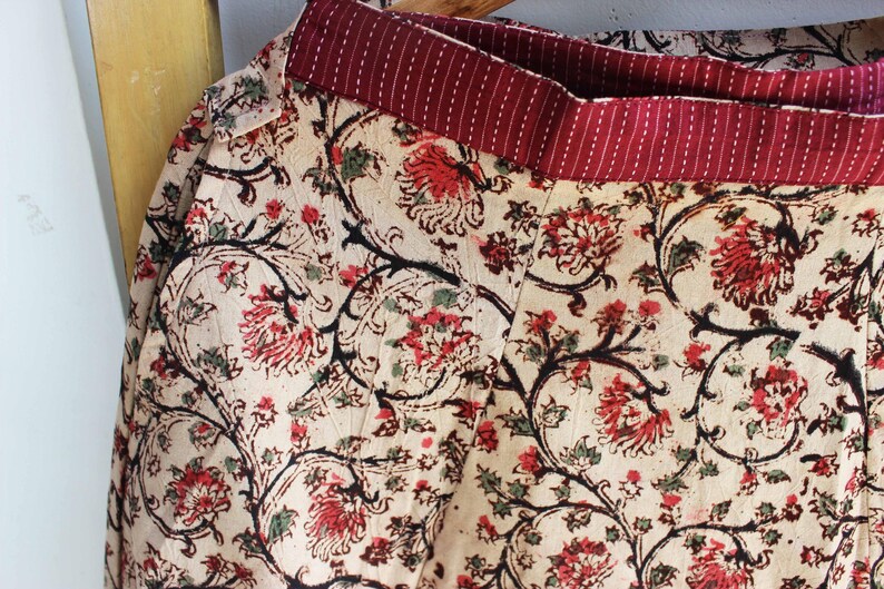 Op de afbeelding: Een rok met een cr&egrave;mekleurige basis en een herhalend bloemenpatroon in tinten rood, zwart en groen. Het kledingstuk heeft een brede, donkerrode tailleband met dunne, verticale witte strepen en zichtbare stiksels over de gehele stof.