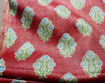 May include: Close-up of a red fabric with a repeating pattern of stylized floral designs. Each design features a green base, white outline, and blue flower clusters. The fabric has a textured appearance.