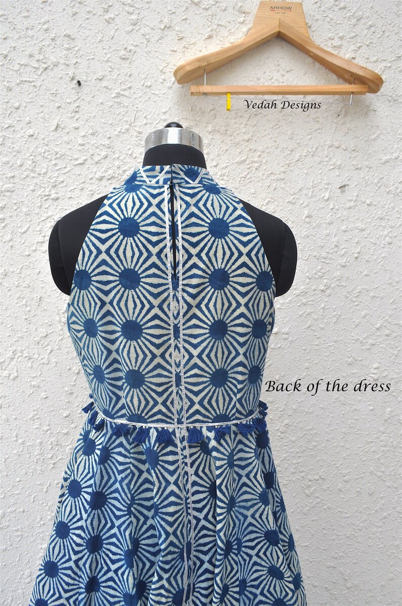 May include: A sleeveless dress featuring a blue and white geometric sunburst pattern, a high neckline, and a vertical zipper closure. The waist is accented with a row of small blue tassels and silver trim. The text Vedah Designs and Back of the dress appear.