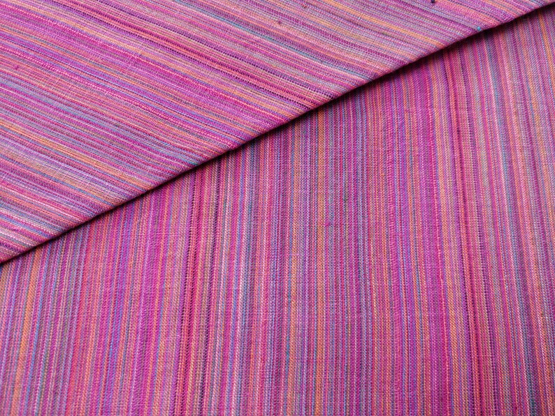 Puede incluir: Primer plano de una tela de rayas rosa y p&uacute;rpura con un brillo sutil. La tela est&aacute; doblada por la mitad, creando una l&iacute;nea diagonal a trav&eacute;s de la imagen.