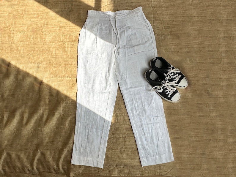 May include: A pair of white trousers with an elasticated waistband and a drawstring closure, featuring a straight-leg cut. Beside the trousers sits a pair of black and white low-top sneakers with white laces and rubber toe caps.